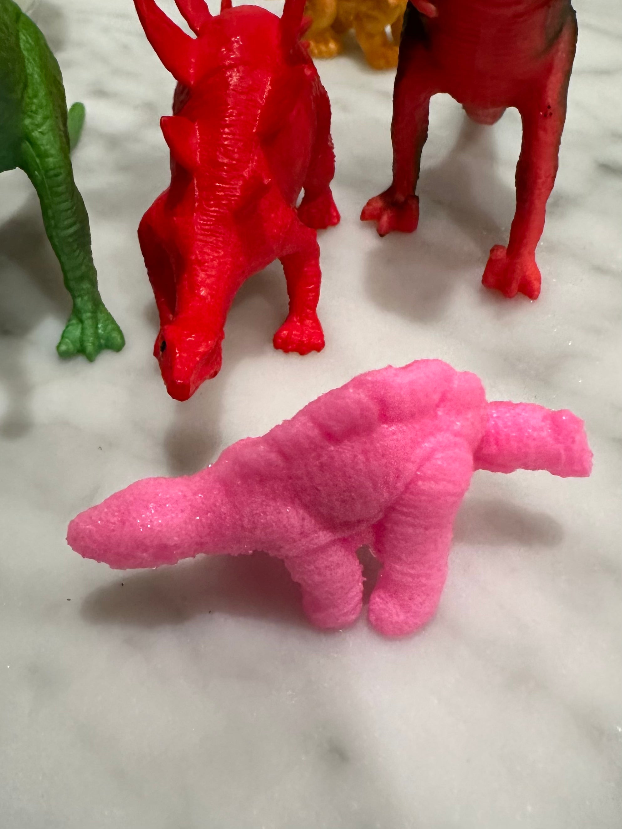 Pink toy dinosaur with red and green dinosaurs in the background, part of a colorful dinosaur playset for kids.