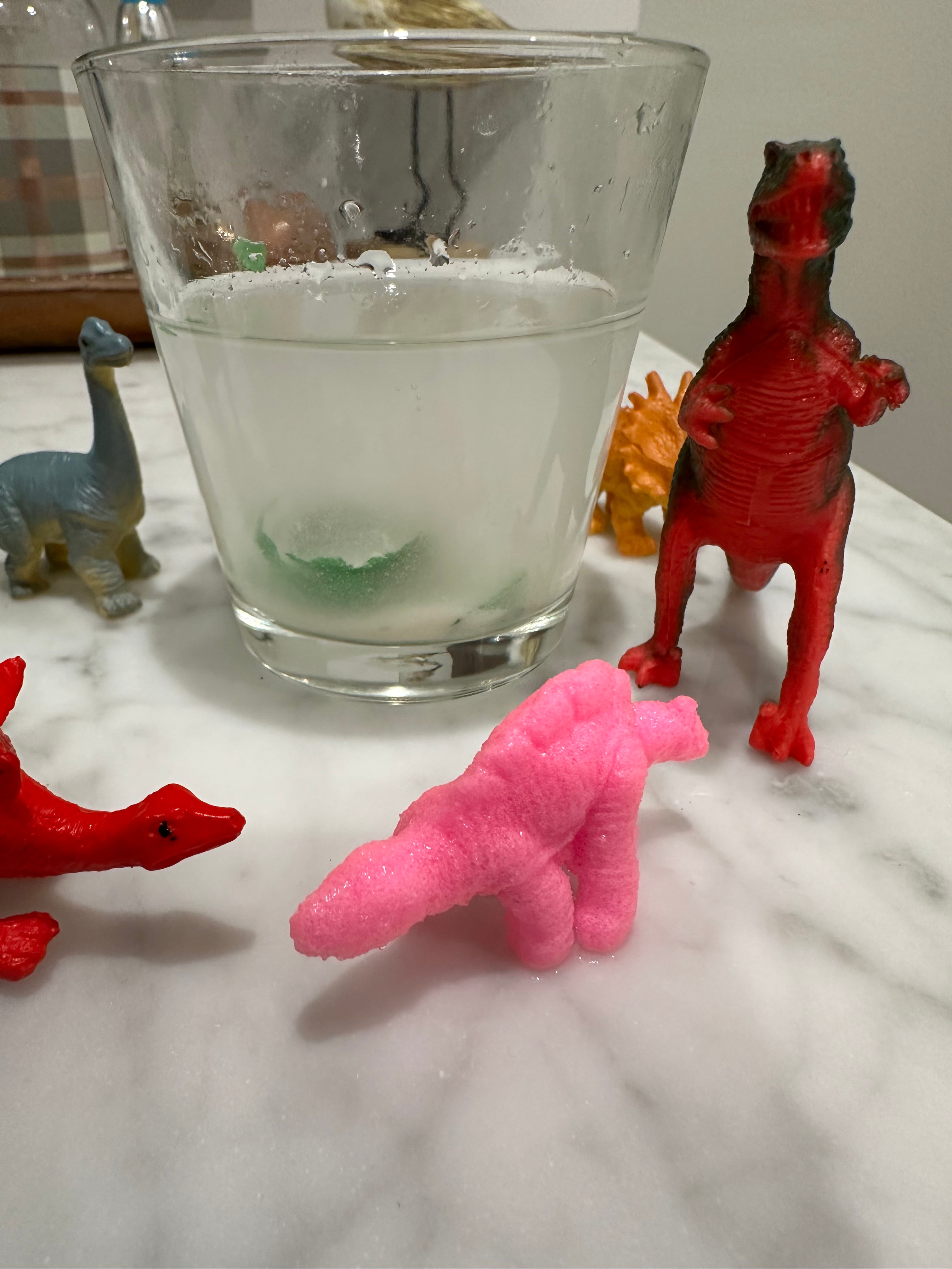 Colorful toy dinosaurs around a glass of water on a marble table, fun educational sensory activity for kids.